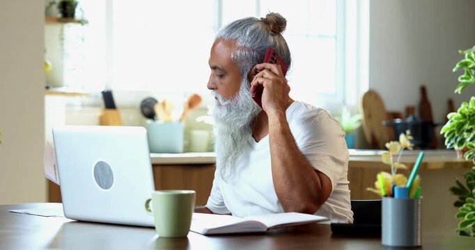 Share trading from home, Indian mid age stylish man with long white beard and hair using smartphone and laptop reviewing company report, writing notes in diary during morning trading session