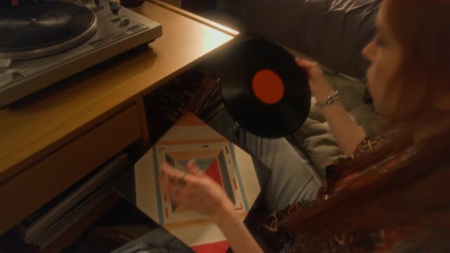 Over shoulder shot of young woman choosing music records and taking out vintage record from sleeve with soft warm light reflecting in black shiny surface