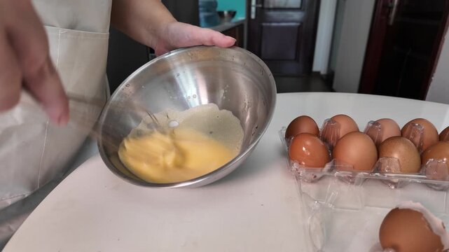 A woman is beating eggs with a whisk in a metal bowl on the kitchen table