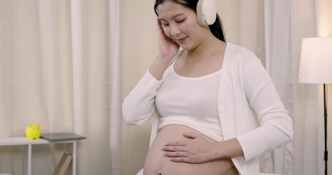 Asian pregnant woman gently touching headphones with one hand while resting other hand on belly, enjoying bonding with unborn baby through music during third trimester in quiet home environment