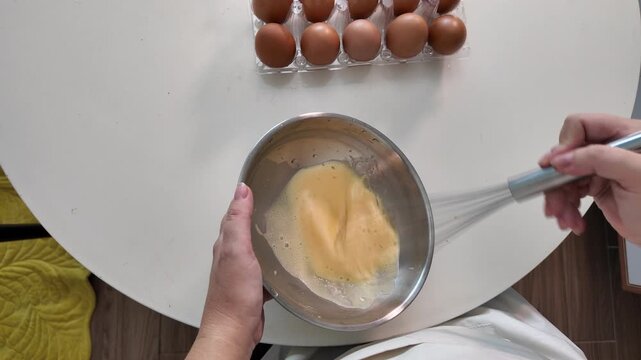 Woman is beating eggs with a whisk in a metal bowl on the kitchen table
