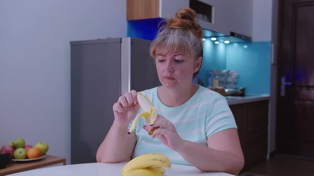 A woman peels and eats a banana sitting at a table , kitchen background