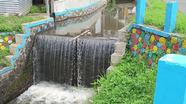 A steady stream of water flows over a small dam in an irrigation canal. The surrounding stone walls are painted with bright, colorful circular patterns.