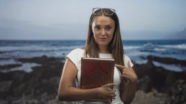 Woman holding ornate red book points finger to chest in studio with sunglasses on head and embossed hardcover by ocean coast backdrop; self identity confidence.