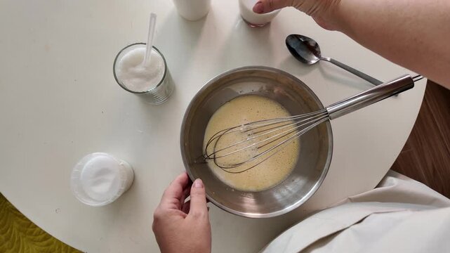 The hands of a woman who adds flour and milk to the beaten eggs for cooking, whips them with a whisk