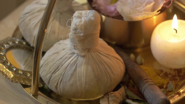 Arrangement of herbal massage compress balls on a golden tray next to a burning candle. Spa setting for Ayurvedic pinda sweda treatment focusing on relaxation, heat therapy, and natural healing.