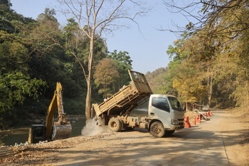 Dump truck unloading gravel for road repair at rural construction site. An excavator and heavy machinery diligently work on infrastructure development in mountain area © CoreRock
