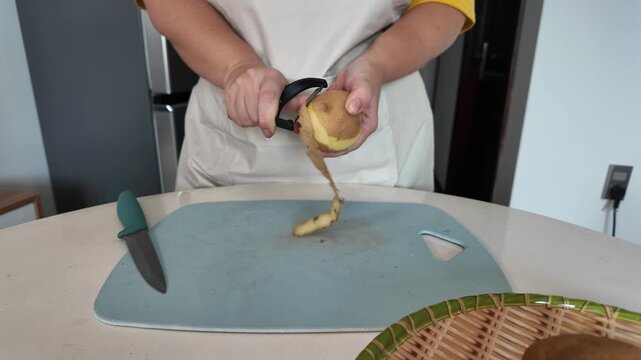 The hands of a woman who, in an apron, peels potatoes with a vegetable peeler while standing