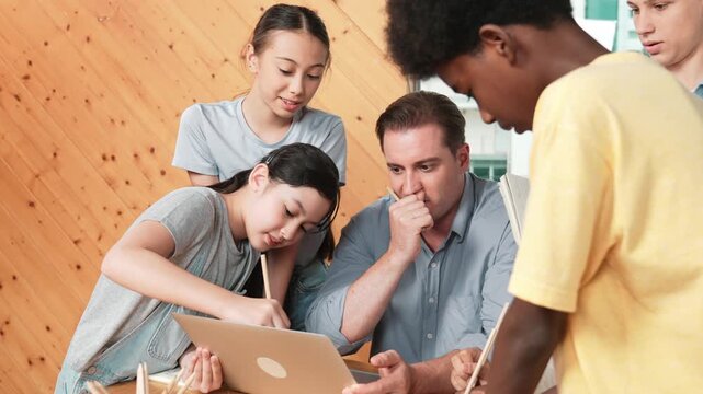Caucasian teacher giving comment or advise student while smart bot pointing at laptop screen at classroom. Diverse student taking a note while teacher teach lesson while using computer. Edification
