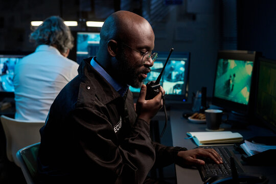 Blackman wearing security uniform monitoring surveillance footage and speaking into walkie talkie while sitting at desk in control room with multiple monitors, female colleague visible in background