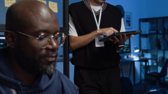 Close-up rack focus shot of bearded African American young IT specialist wearing glasses working in modern office, product manager with badge using digital tablet in background