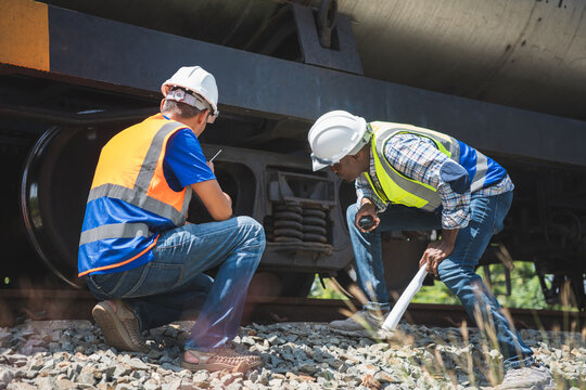 Diverse railway engineers inspecting train wheels and undercarriage at a construction site, Maintenance team with walkie-talkie checking oil tanker train components for logistics