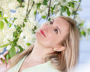 Fototapeta premium Real people, simple authentic natural beauty of a Caucasian blonde woman with candid smile wearing light cotton shirt. Scene in a spring park at the blooming choke-cherry tree.