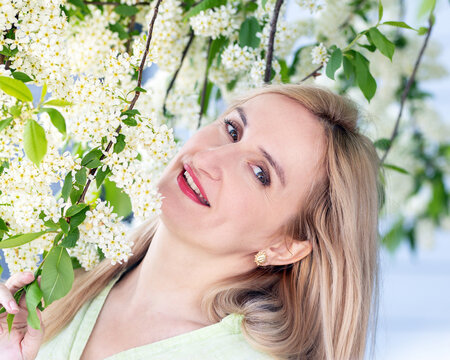 Real people, simple authentic natural beauty of a Caucasian blonde woman with candid smile wearing light cotton shirt. Scene in a spring park at the blooming choke-cherry tree.