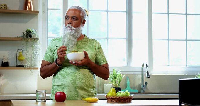 Indian mid age man eating poha while standing in home kitchen in the morning, stylish bearded male holding bowl with healthy breakfast and enjoying traditional Indian meal with spoon