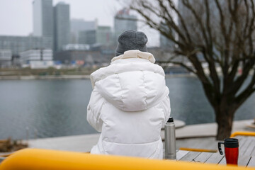 Young woman in a white puffer jacket sits on a bench, overlooking a foggy city waterfront. Concept of an urban winter activity © Volha