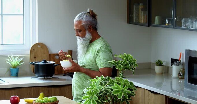 Indian mid age man eating poha while standing in home kitchen in the morning, stylish bearded male holding bowl with healthy breakfast and enjoying traditional Indian meal with spoon