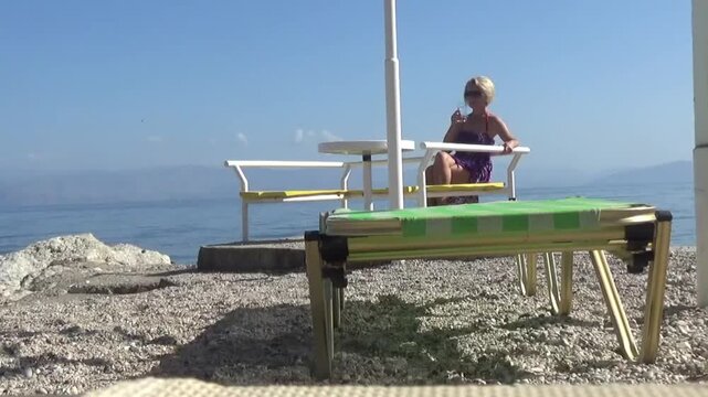 A young blonde in a pareo sits at a table on the beach, drinking water alone. Taking a break from swimming and relaxing