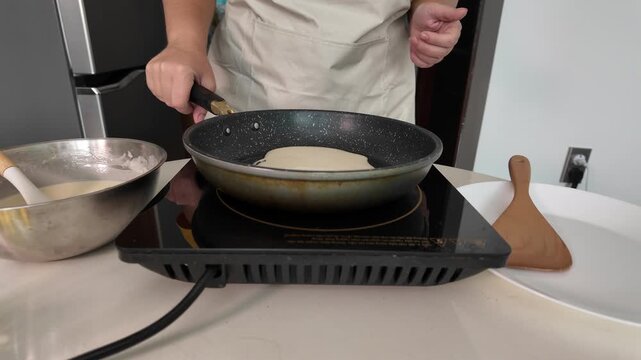 Woman is frying pancakes at home in the kitchen on an induction hob. The woman distributes the dough evenly over the hot pan.