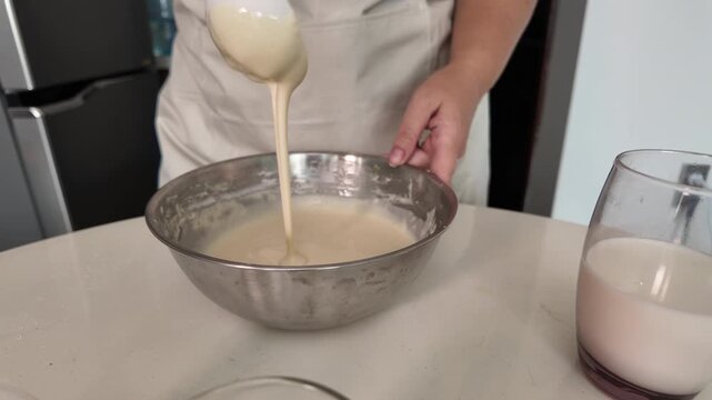 A woman in an apron, using a ladle, is stirring pancake batter in a metal bowl