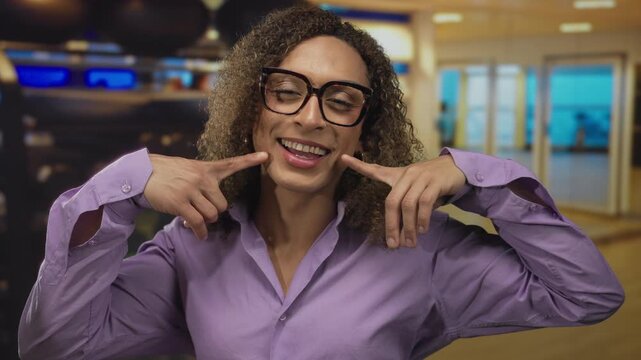 Grinning transgender woman in purple shirt finger pointing to cheek in indoor gym with mirrored wall; joy pride.
