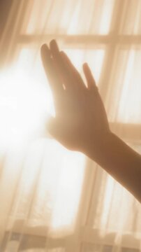Vertical shot of anonymous girl waving hand against bright sun light shining through curtains indoors with lens flare effect