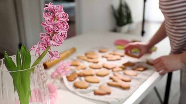 A person prepares Easter cookies in a bright kitchen. They use cookie cutters to shape dough and decorate a table with flowers. The activity is fun and engaging, focusing on creativity.