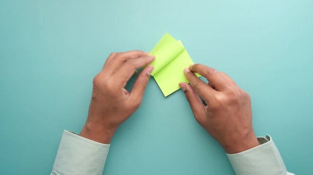 A pair of hands neatly tears a green sticky note in half, creating two smaller square pieces of paper, shot from an overhead perspective on a teal surface