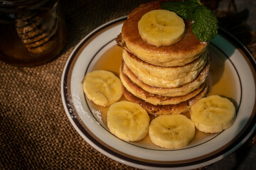 Stack of fluffy homemade banana pancakes topped with fresh slices, mint leaf, and golden honey dripping from a wooden dipper. Rustic breakfast concept on burlap background with natural sunlight. © Chayut