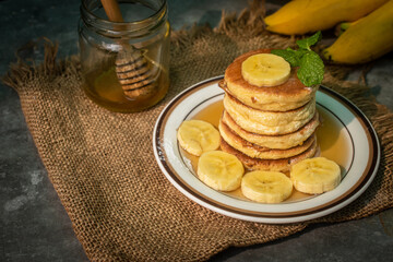 Stack of fluffy homemade banana pancakes topped with fresh slices, mint leaf, and golden honey dripping from a wooden dipper. Rustic breakfast concept on burlap background with natural sunlight. © Chayut
