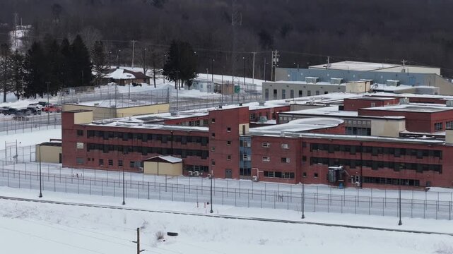 Aerial view of brick Federal Correctional Institution complex in Loretto, Pennsylvania, surrounded by security fencing and snow-covered grounds during winter, with wooded hills in background.Wide shot