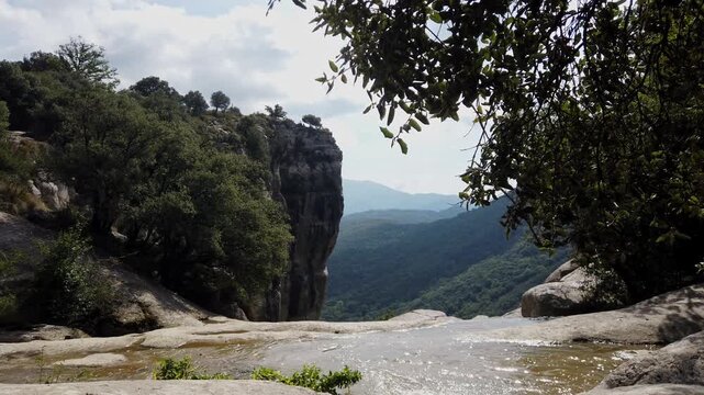 Scenic view of a creek flowing over rocks towards the edge of a waterfall, with a breathtaking panoramic view of the green valley and mountains in Rupit, Catalonia, on a sunny day