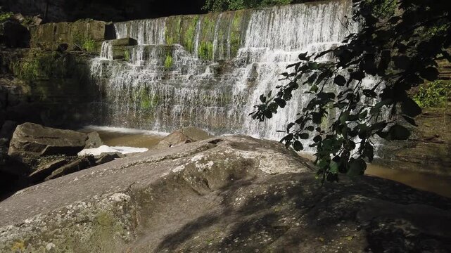 Scenic view of a powerful waterfall cascading down rocky cliffs in a green forest, creating a serene and wild natural landscape in Rupit, Catalonia, Spain during a sunny day