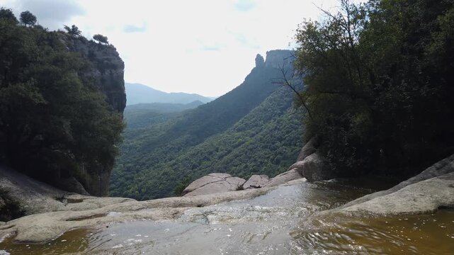 Stunning first-person perspective of a mountain stream flowing towards the edge of a high cliff, overlooking a vast green valley in Rupit, Catalonia, on a summer day with a cloudy sky