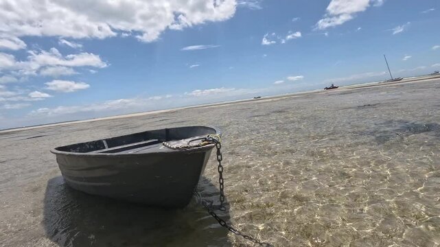 A dingy punt rests in shallow water under a bright sky. Additional boats are visible in the distant background. The setting is coastal and outdoor.