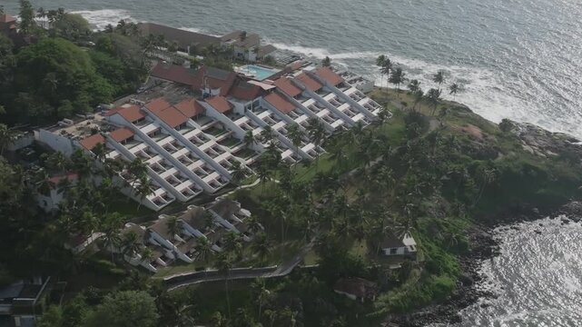 Aerial showcases Charles Correa's iconic tiered Kerala vernacular design: white walls, red roofs cascading down cliffs amid palms to Arabian Sea waves.