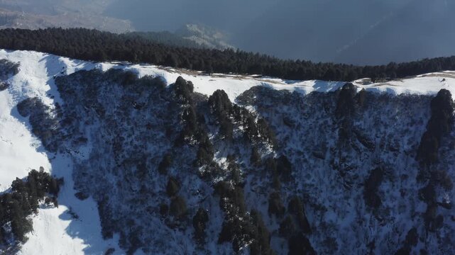 Gentle drone pan across dark pine woods with snow rims unveils layered hazy mountains in golden light at high-altitude Jalori Pass.