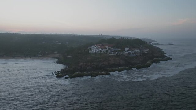 Aerial dawn view of The Leela resort on palm-covered cliff rising from ocean waves, bathed in soft pink-orange sunrise light with hazy skies and distant coastline.