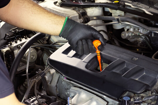 Manual Engine Oil Check, A mechanic's gloved hand pulls the orange dipstick from a black engine cover to inspect motor oil levels and quality.