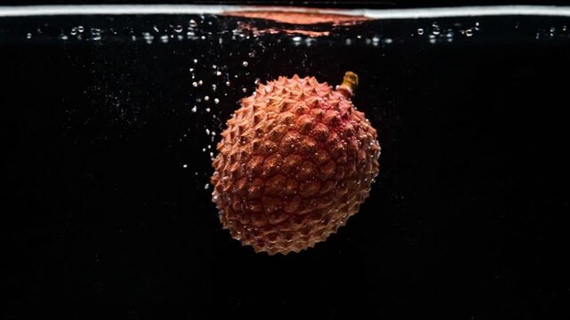 A vibrant red lychee fruit with textured skin plunges into clear water, creating bubbles against a dark background.