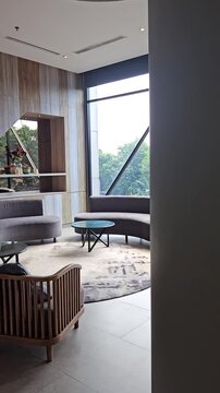 Modern luxury hotel lobby lounge with stylish curved grey sofas, elegant wooden chairs, marble floors, and large glass windows offering a bright atmosphere and scenic green outdoor views for guests
