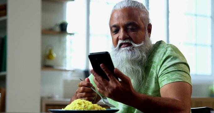 Indian mid age man watching smartphone reels while eating at home, stylish bearded male enjoying poha at breakfast table and streaming web series or comedy videos in modern kitchen lifestyle