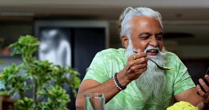 Indian mid age man watching smartphone reels while eating at home, stylish bearded male enjoying poha at breakfast table and streaming web series or comedy videos in modern kitchen lifestyle