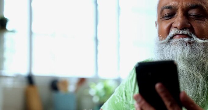 Indian mid age man watching smartphone reels while eating at home, stylish bearded male enjoying poha at breakfast table and streaming web series or comedy videos in modern kitchen lifestyle