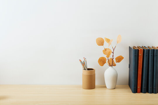 Minimalist home office desk setup with notebooks, a white vase with dried leaves, and stationery on a light surface