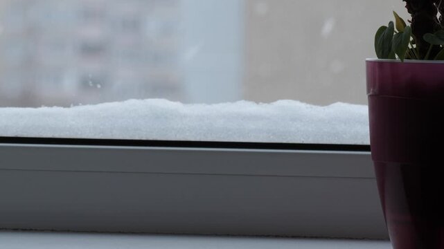 Snowdrift on a window frame next to a pink flower pot. View of the winter city through a snowy window during a snowfall