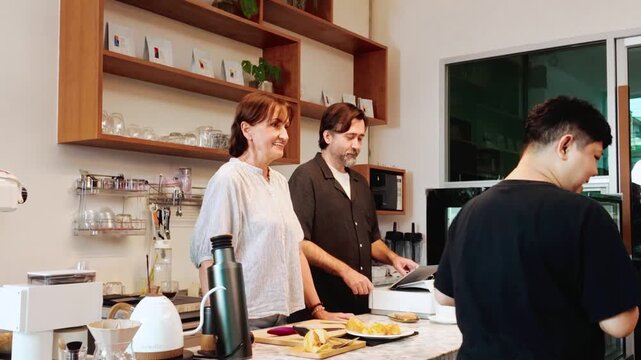 Baristas serve pastries and coffee to a customer at a cozy cafe counter, with shelves of cups and plants behind and a display case beside them.