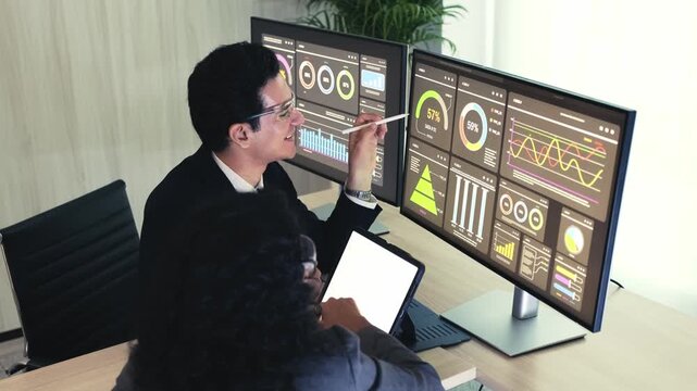 Two professionals review analytics dashboards on dual monitors in a modern office, discussing performance metrics and trends.