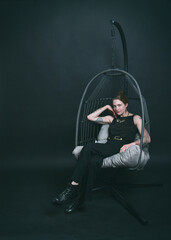 Naklejka premium Ganzkörperportrait einer Frau im Hängesessel vor dunklem Studiohintergrund. Lässige Pose, moderne Ästhetik und stimmungsvolles Licht – Fashion trifft Interior-Design.