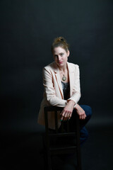 Naklejka premium Stimmungsvolles Frauenportrait im Low-Key-Licht. Selbstbewusstes Model mit Blazer auf Stuhl vor dunklem Hintergrund – modernes Business- und Charakterportrait mit Tiefe.